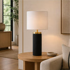 A modern table lamp with a black ribbed cylindrical base and a white shade, on a round wooden side table