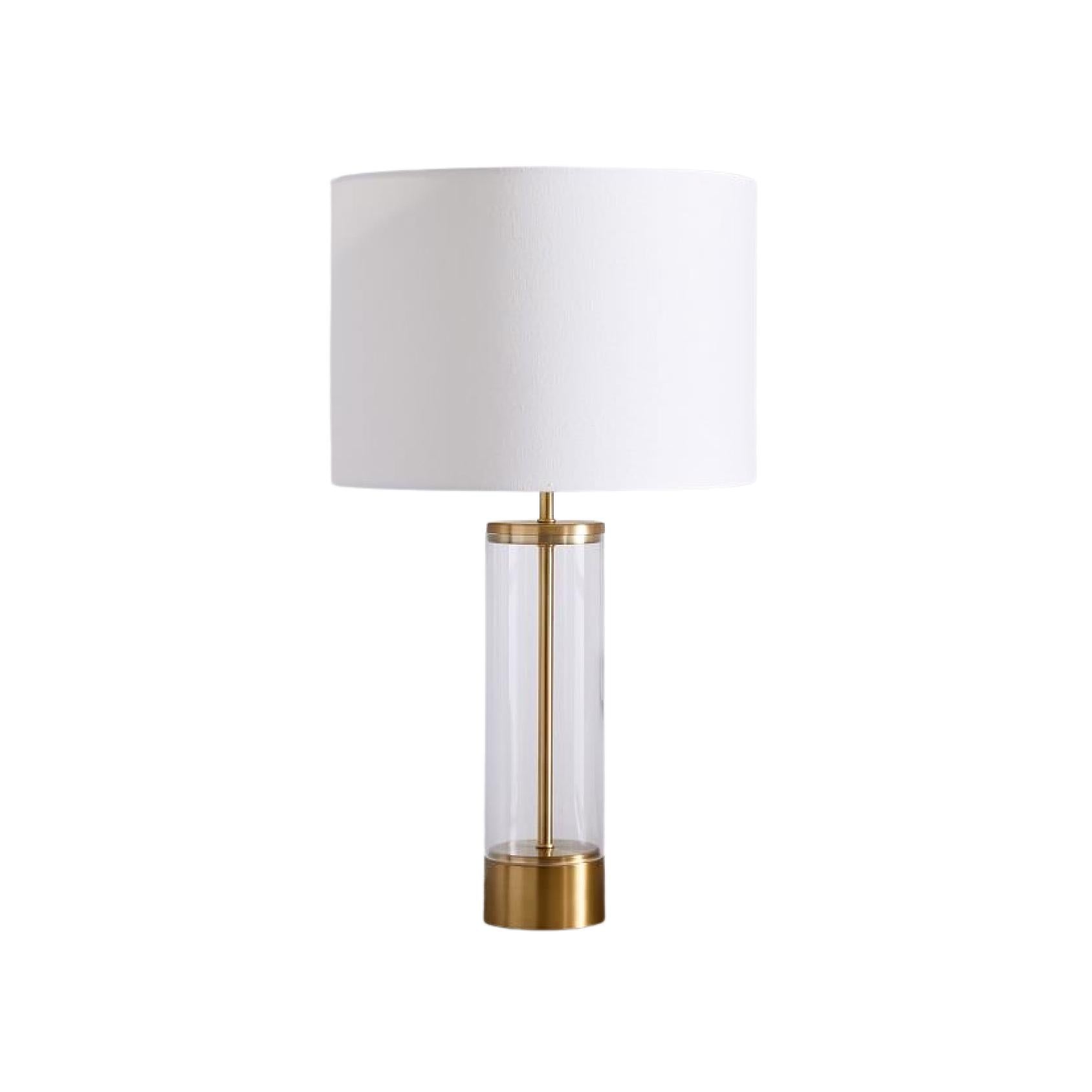 Table lamp with a white shade, a clear glass cylindrical body and gold detailing