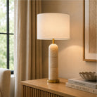 A table lamp with a cylindrical beige stone base and a white shade, set on a wooden console table