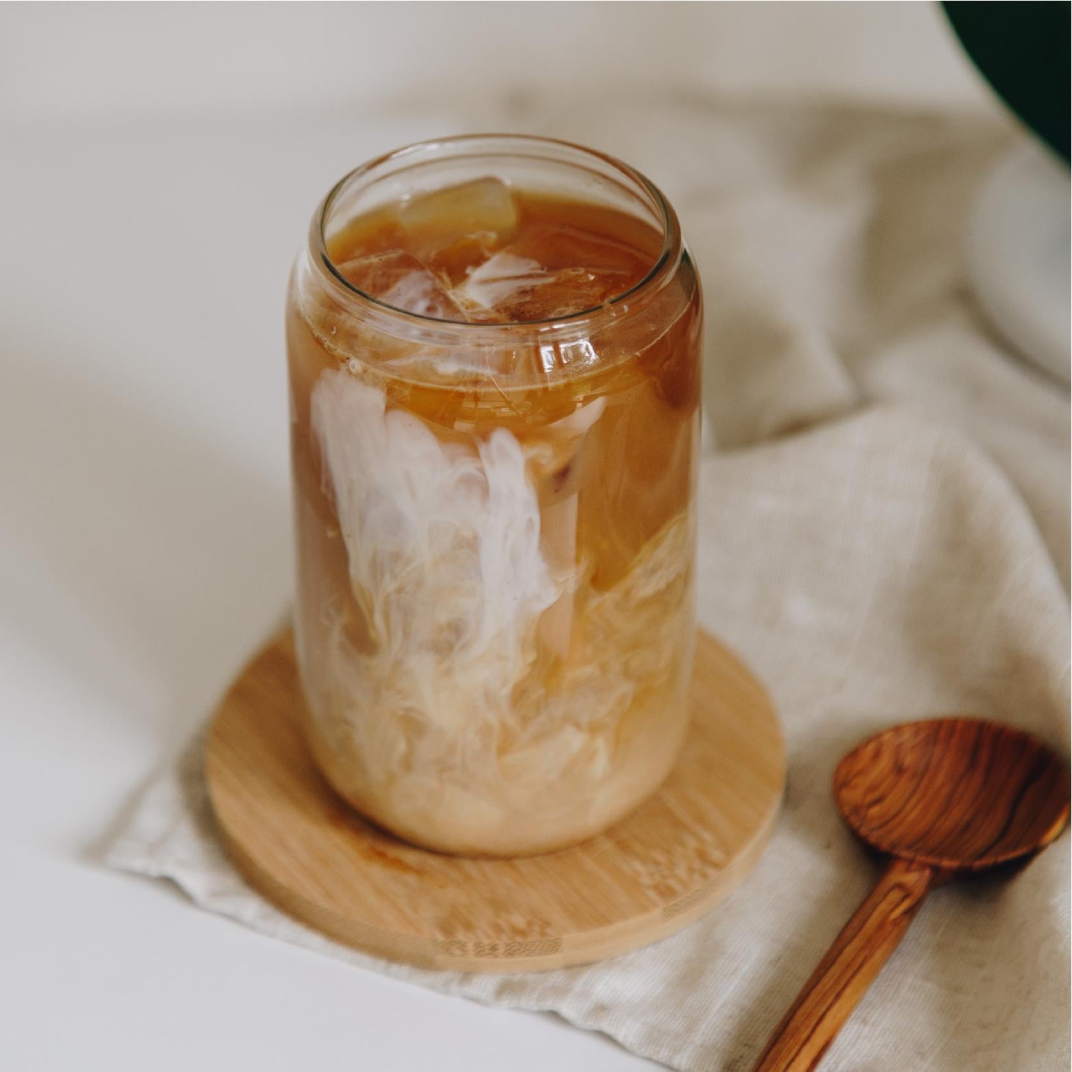 Glass can shaped drinking glass with glass straw on a bamboo coaster, shown with iced coffee